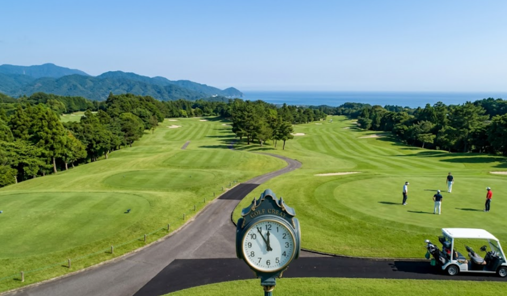 広大な日本のゴルフコースの全景。青空の下、グリーンのアンジュレーション、遠くの山々、そしてプレーするゴルファーが見え、18ホールの広さとプレーの広がりを感じさせる。