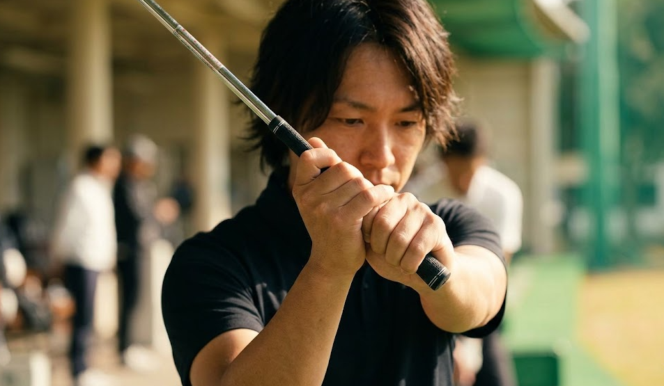 ゴルフ練習場で細いグリップのクラブを握り、フェースターンを意識する日本人ゴルファー。指先でしっかりとホールドし、手首の返しやすさが強調されている。