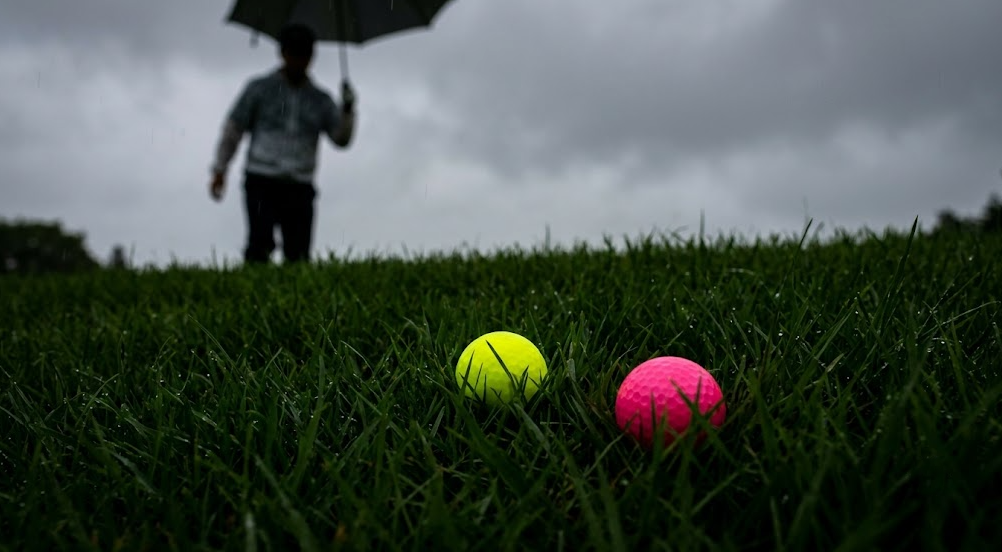 曇り空の下、雨に濡れた深い緑色のラフに深く沈んだ蛍光イエローと蛍光ピンクのゴルフボール。